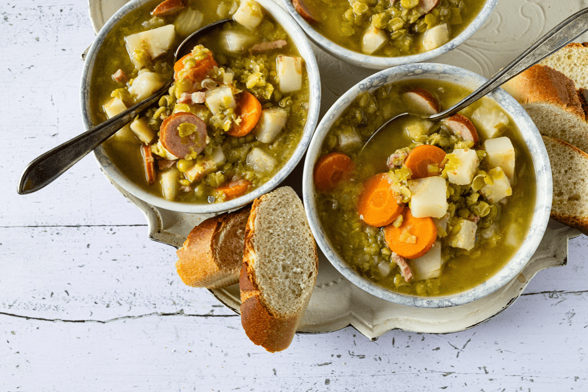 Drie kommen met oma's erwtensoep (snert), gevuld met wortel, aardappel en plakjes rookworst, geserveerd met brood.