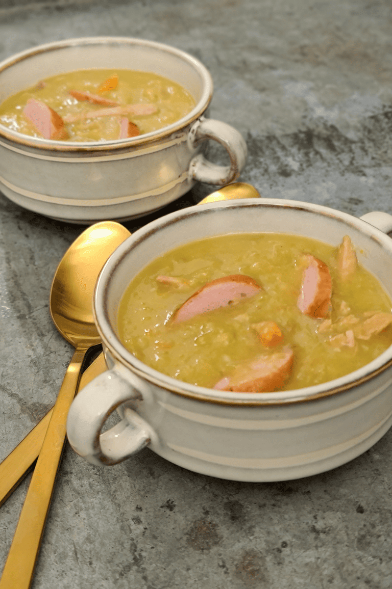twee kommen met ouderwetse erwtensoep en stukken rookworst, geserveerd met gouden lepels.