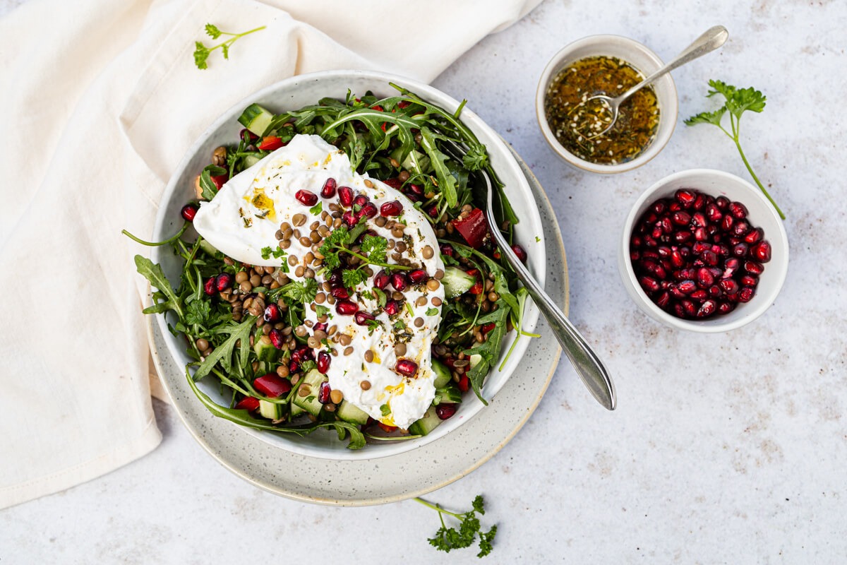Een kom groene linzensalade met burrata, granaatappelpitjes, rucola en een frisse dressing.