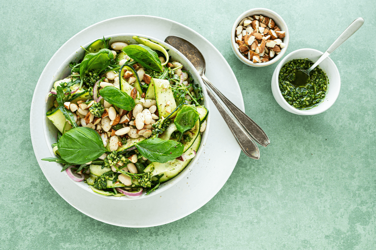 Een witte kom met witte bonensalade met pesto, courgettelinten, amandelen en verse basilicum.