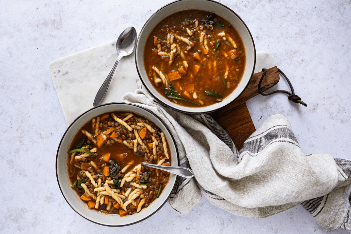 Twee kommen gevulde Italiaanse minestrone soep met pasta, linzen, wortel en spinazie op een houten plank.