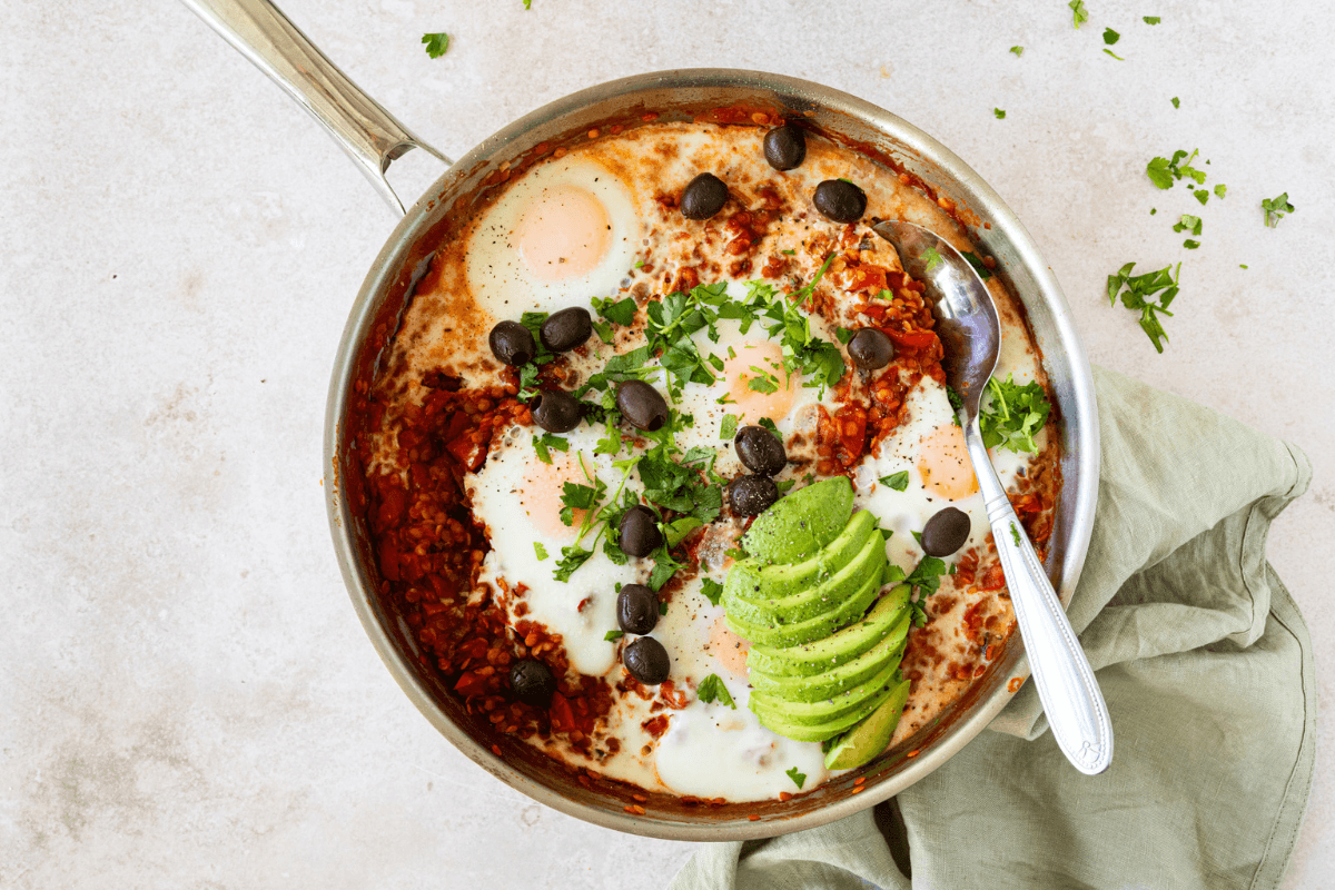 Een pan met shakshuka met linzen, gepocheerde eieren, plakjes avocado, zwarte olijven en verse kruiden.
